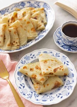 A plate with three folded lefse by a mug of coffee and a platter.