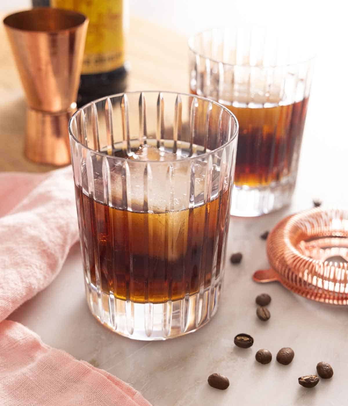 Two Black Russian cocktails with coffee beans scattered by cocktail equipment.