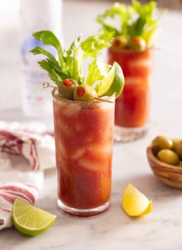 A glass of Bloody Mary with celery, olives, and lime garnish in front of a second one.