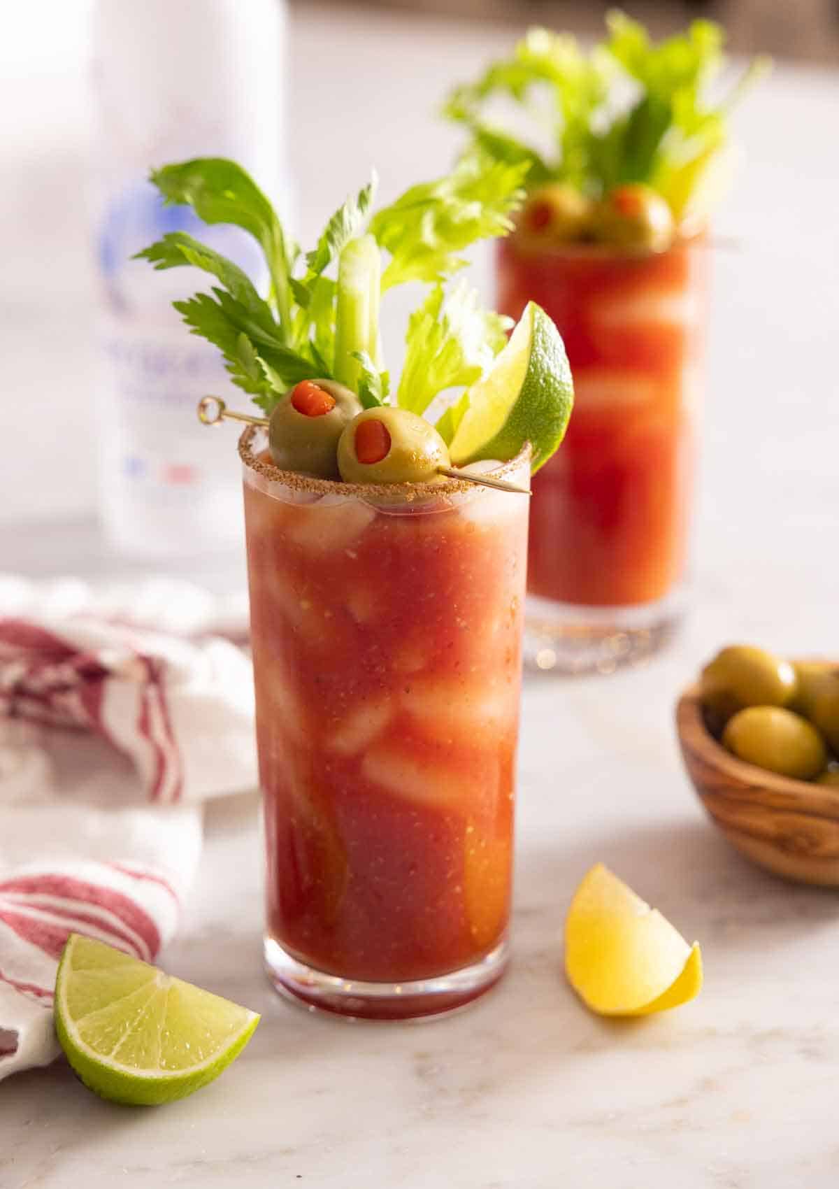 A glass of Bloody Mary with celery, olives, and lime garnish in front of a second one.