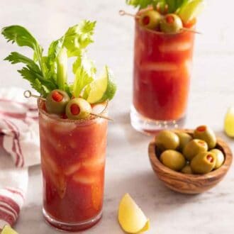 Two glasses of Bloody Marys with lime wedges and a small bowl of olives in between.