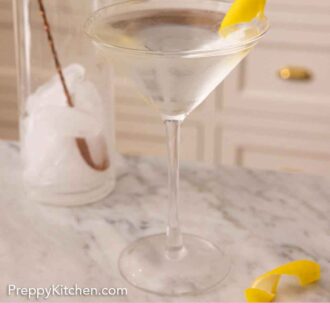 A martini with lemon twist garnish in front of a mixing glass.