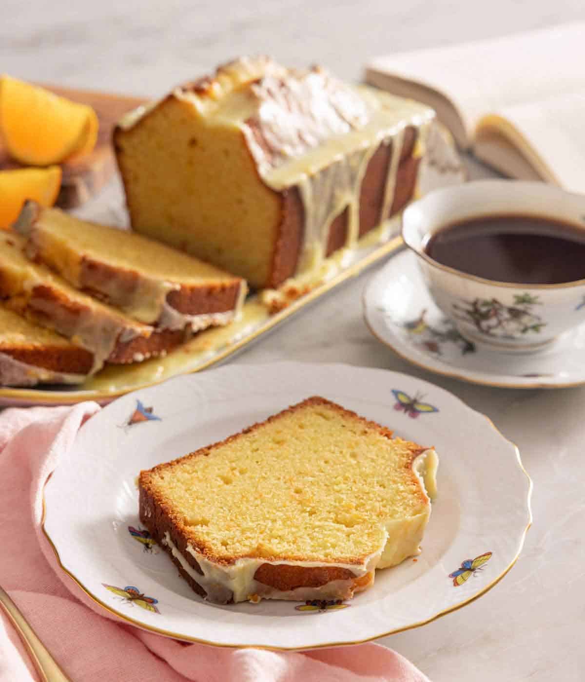 A slice of orange cake in a plate in front of a platter with the rest of the loaf and coffee.