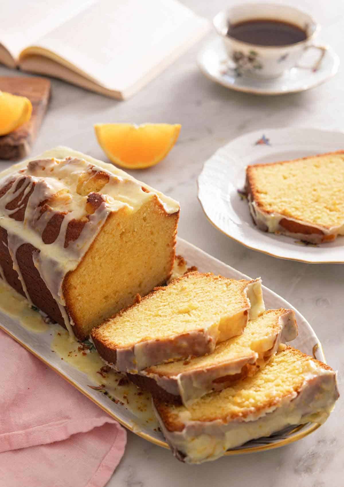 A loaf of orange cake with three slices cut by a mug of coffee and orange wedges.