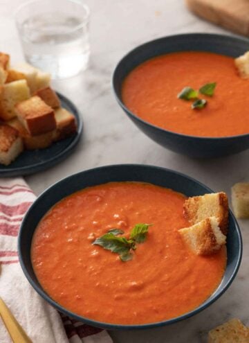 Two bowls of tomato soup with basil and croutons on top.