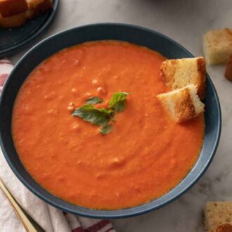 A bowl of tomato soup with basil and croutons scattered around and as garnish.