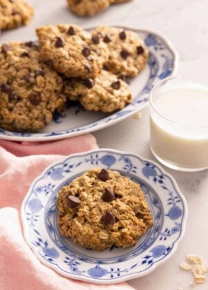 A plate with a banana oatmeal cookie by a glass of milk and a platter with more.