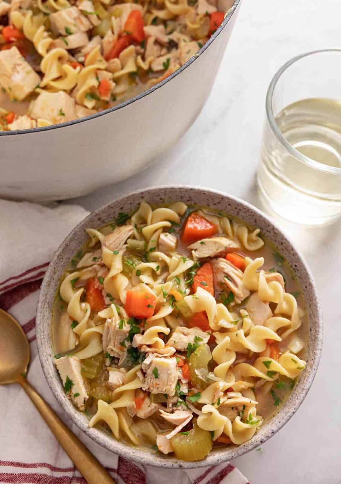 A bowl of chicken noodle soup by a glass of wine.