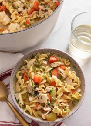 A bowl of chicken noodle soup by a glass of wine.