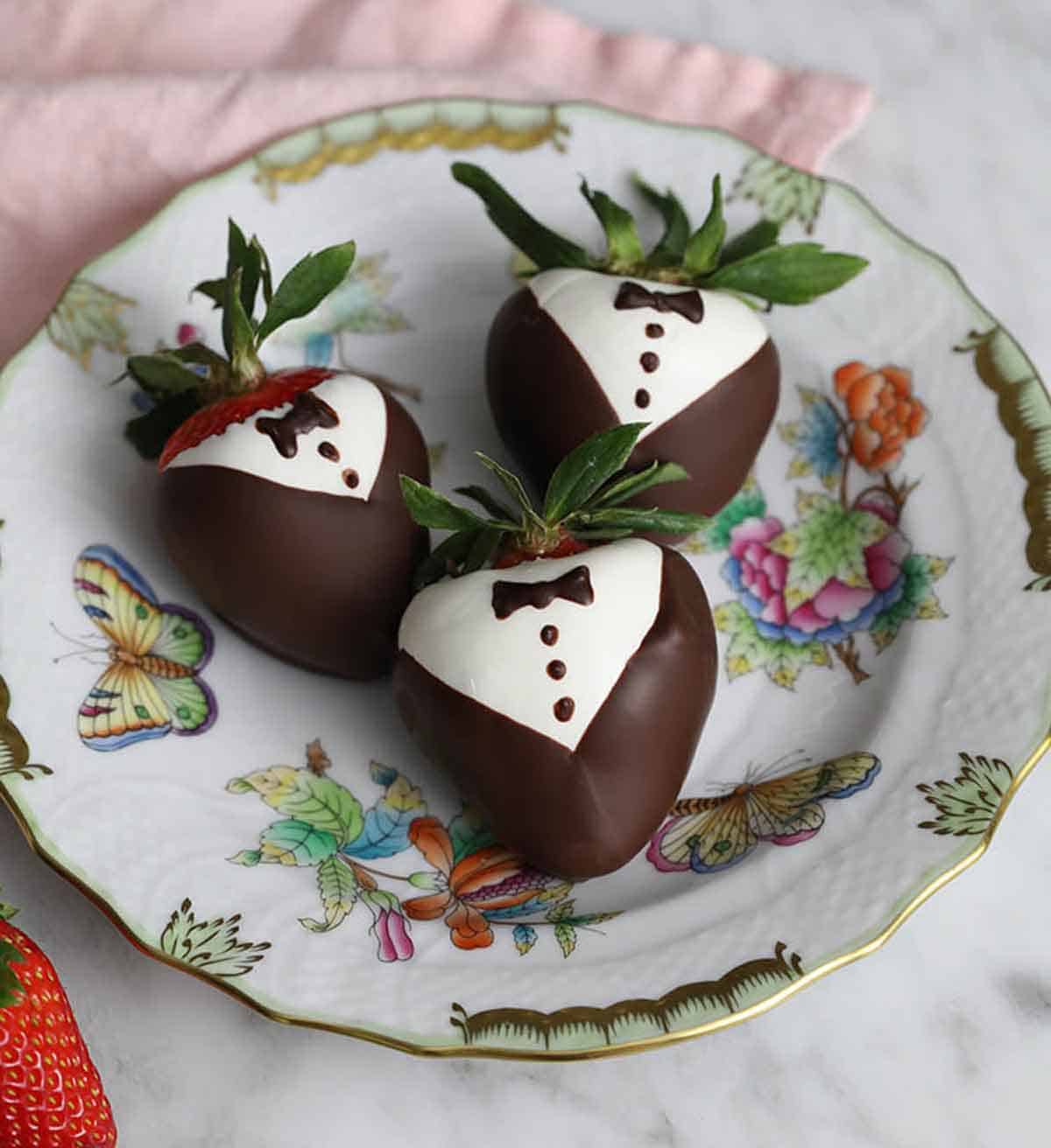 A plate of three chocolate covered strawberries decorated like tuxedos.