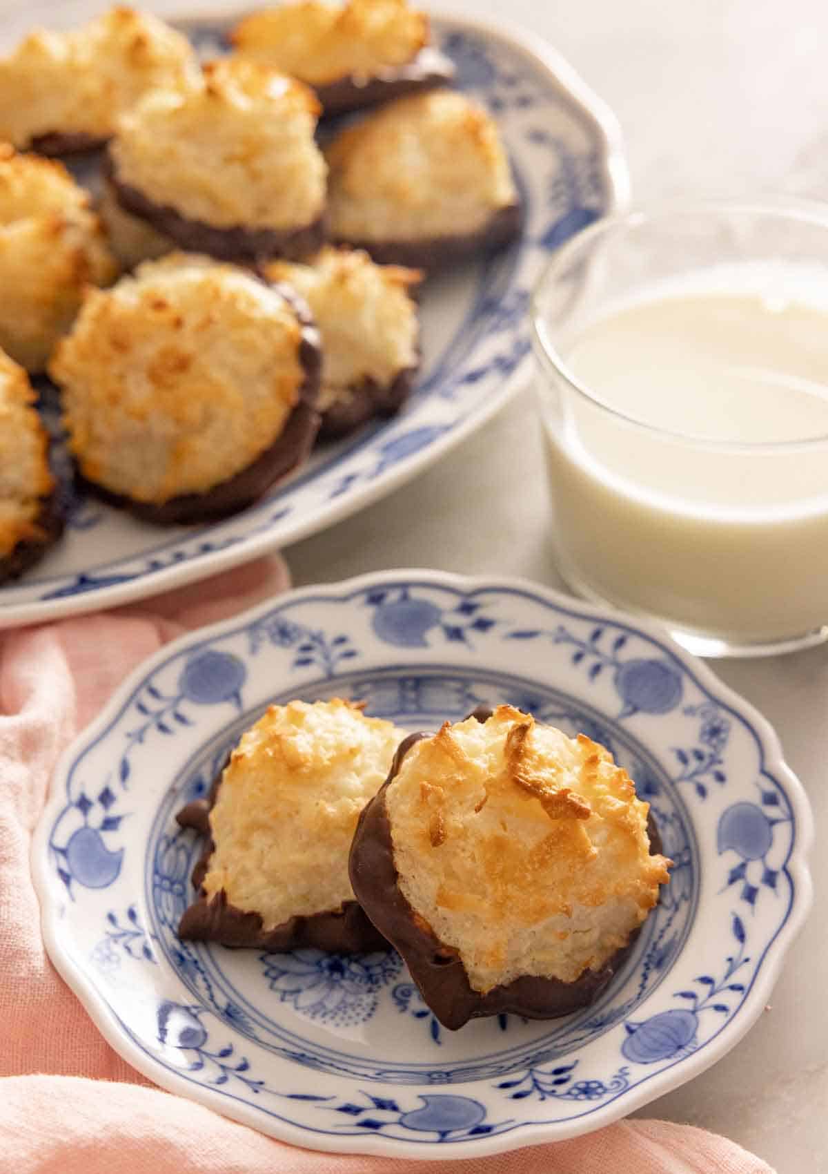A plate with two coconut macaroons in front of a glass of milk and a platter.