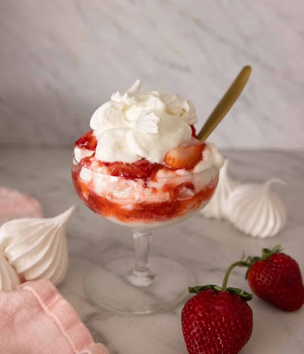A glass of Eton mess with strawberries and meringue cookies scattered around.
