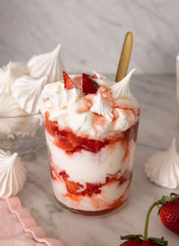 A glass of Eton mess with one in front with strawberries and meringue cookies around it.