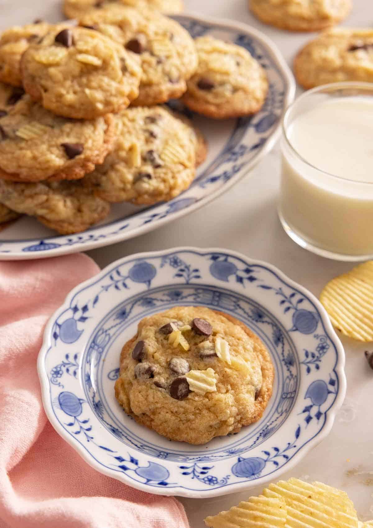 A potato chip cookie on a plate by a glass of milk and more cookies.