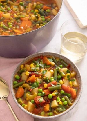 A bowl of vegetable soup by a glass of wine and pot of soup.