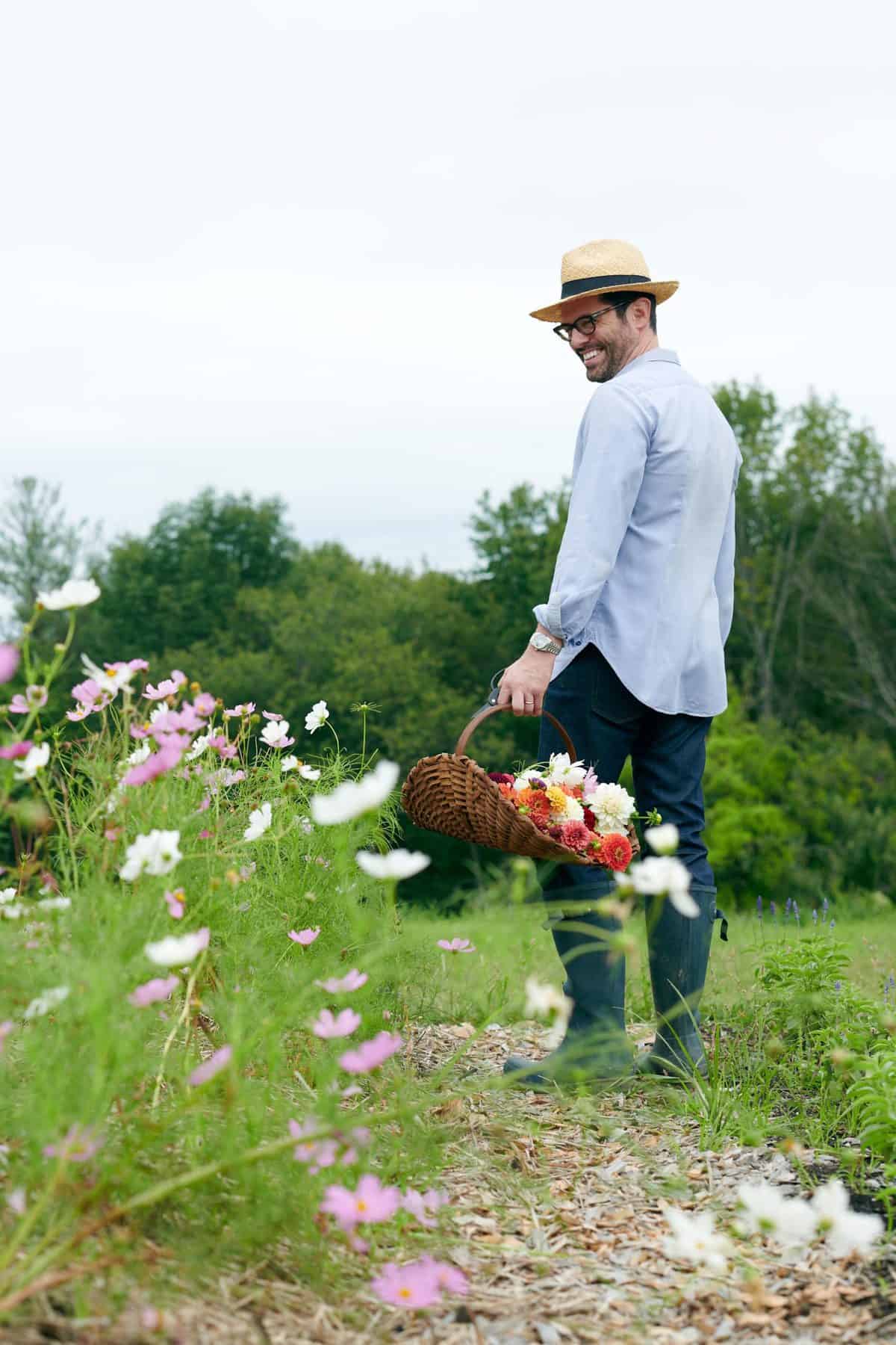John Kanell smiling and walking in a field of flowers while holding a basket of cut flowers.