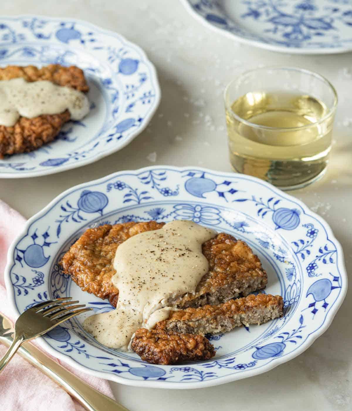 A plate with chicken fried steak sliced, with gravy drizzled on top by a glass of wine.