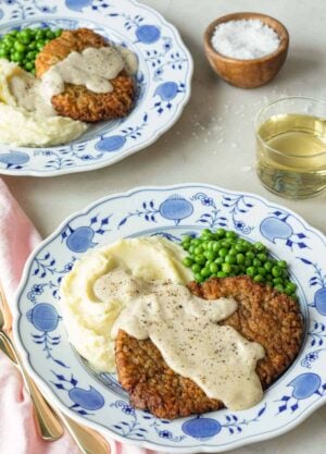 Two plates with chicken fried steak with gravy on top with mashed potatoes and peas.