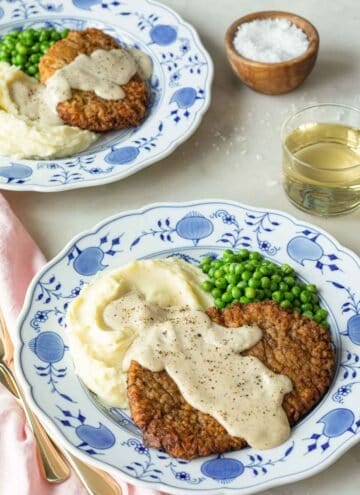 Two plates with chicken fried steak with gravy on top with mashed potatoes and peas.