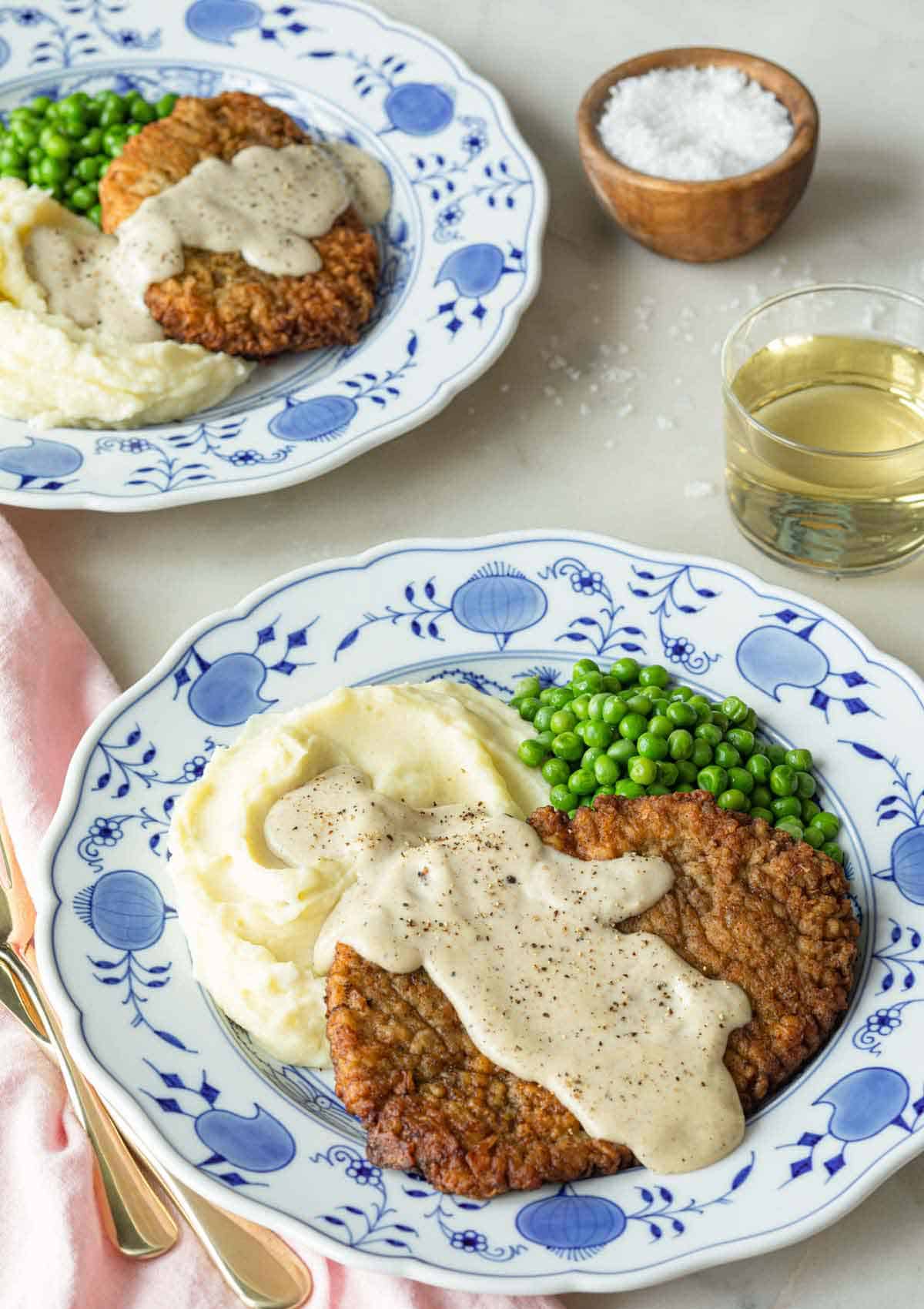 Two plates with chicken fried steak with gravy on top with mashed potatoes and peas.