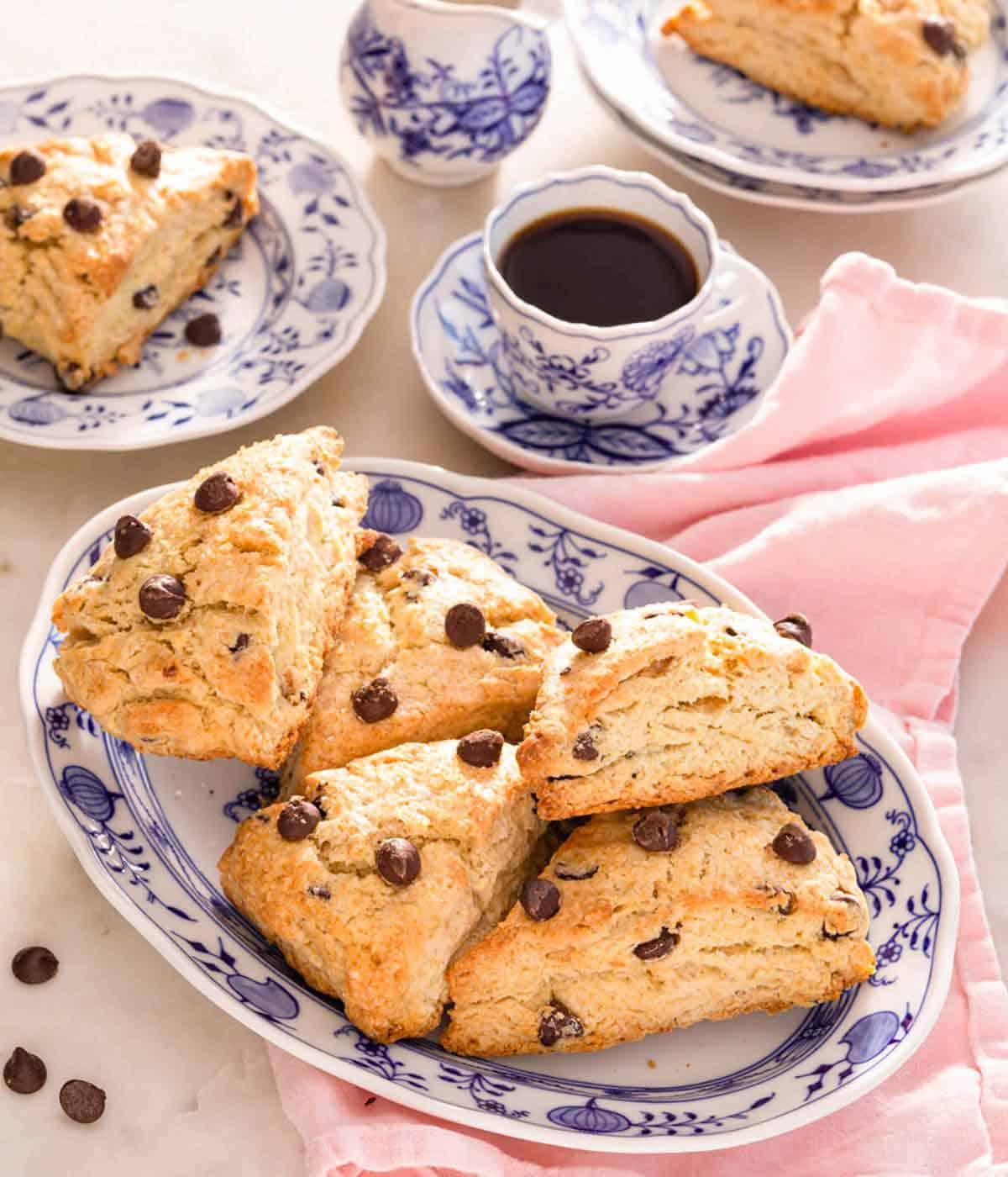 A platter of chocolate chip scones with a cup of coffee and plates with scones behind it.