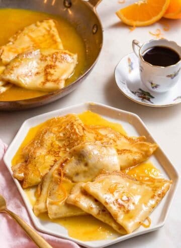 A plate with multiple crepe suzette in front of a cup of coffee and a pan.