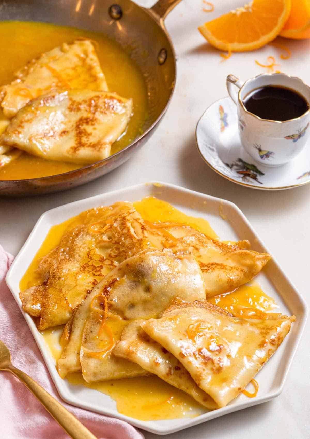 A plate with multiple crepe suzette in front of a cup of coffee and a pan.