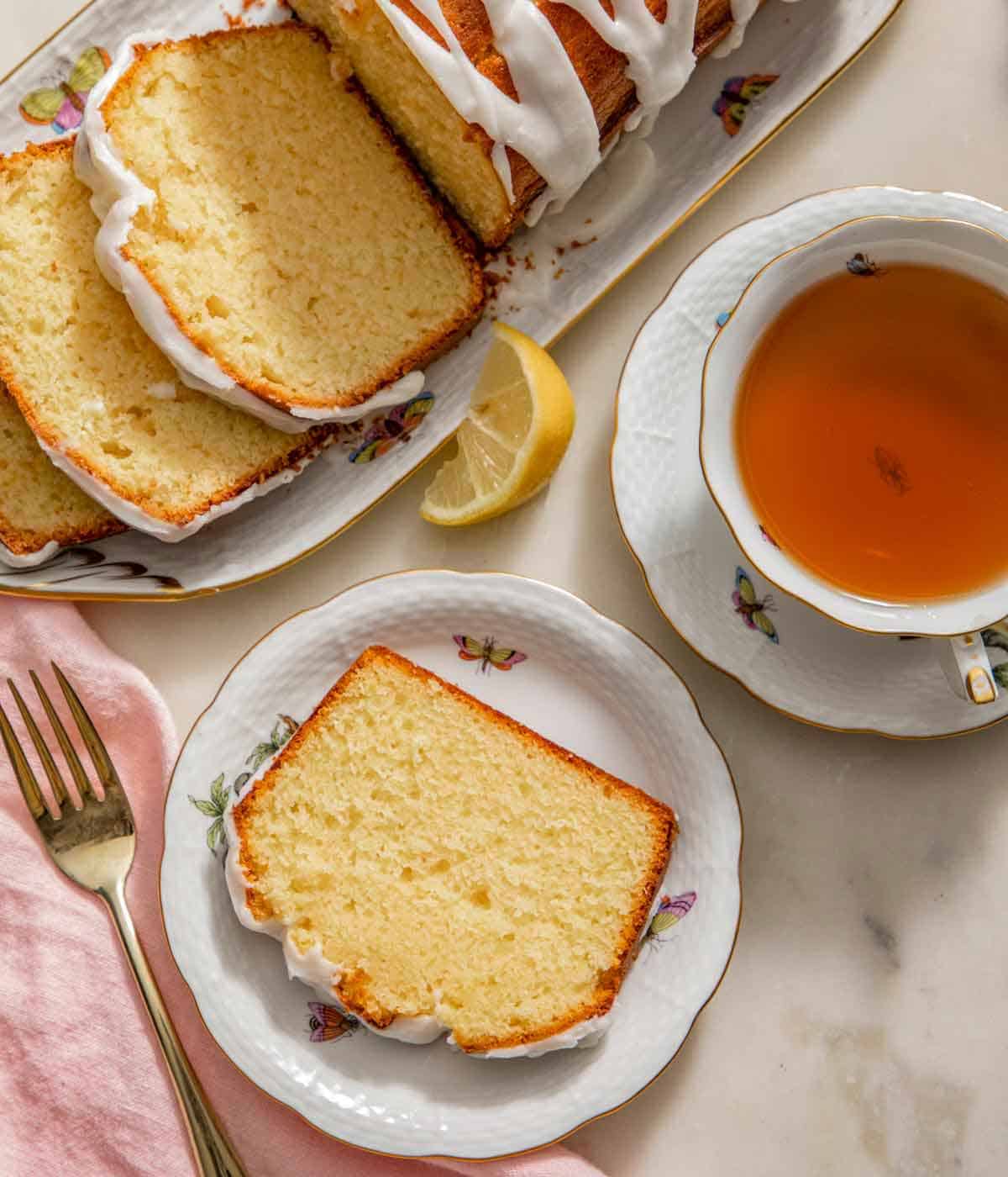 Overhead view of a plate with a slice of lemon pound cake by a cup of tea and the cut loaf.