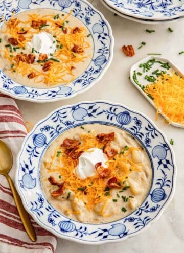 Two shallow bowls of potato soup with shredded cheese, bacon, sour cream, and chives on top.