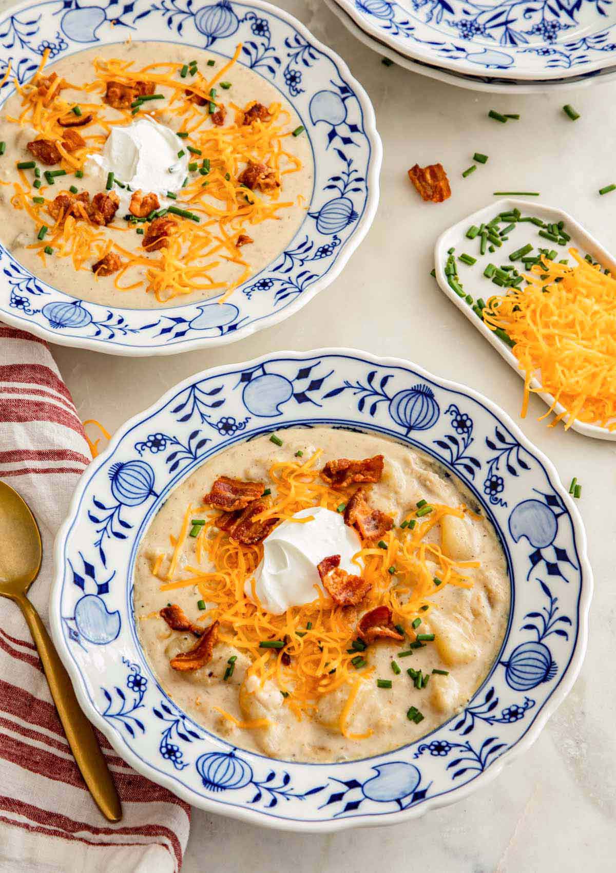 Two shallow bowls of potato soup with shredded cheese, bacon, sour cream, and chives on top.