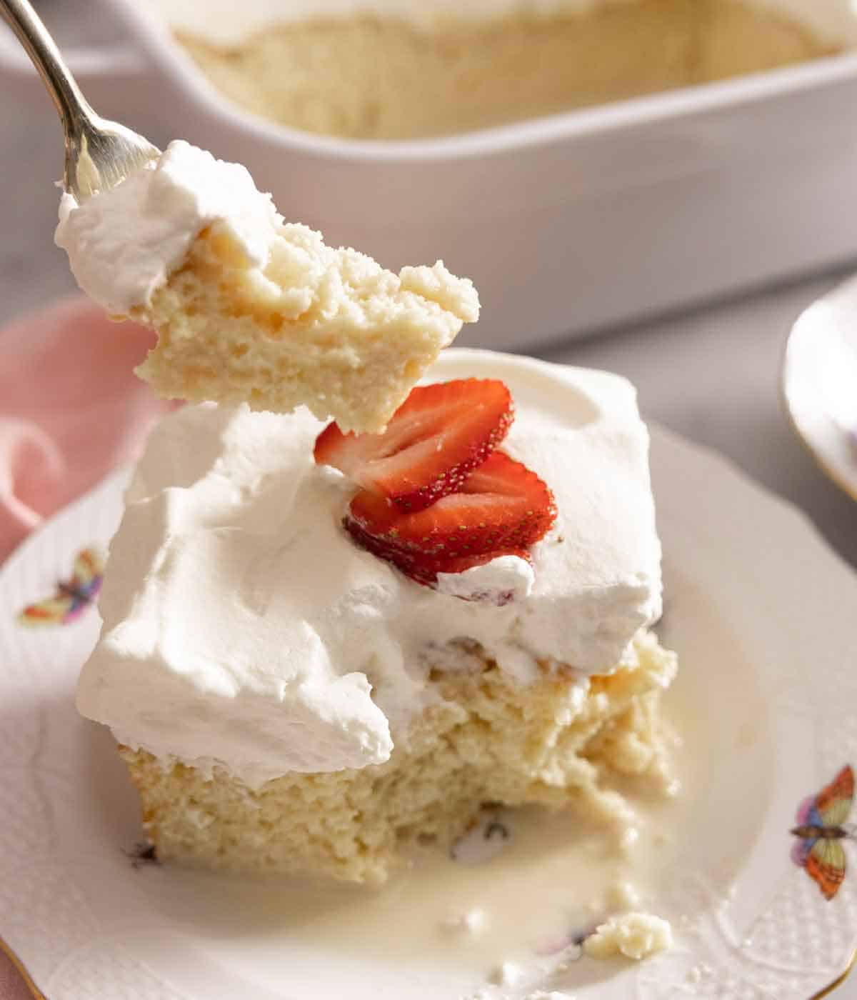 A forkful of tres leches cake lifted from a plate with a slice with strawberry on top.