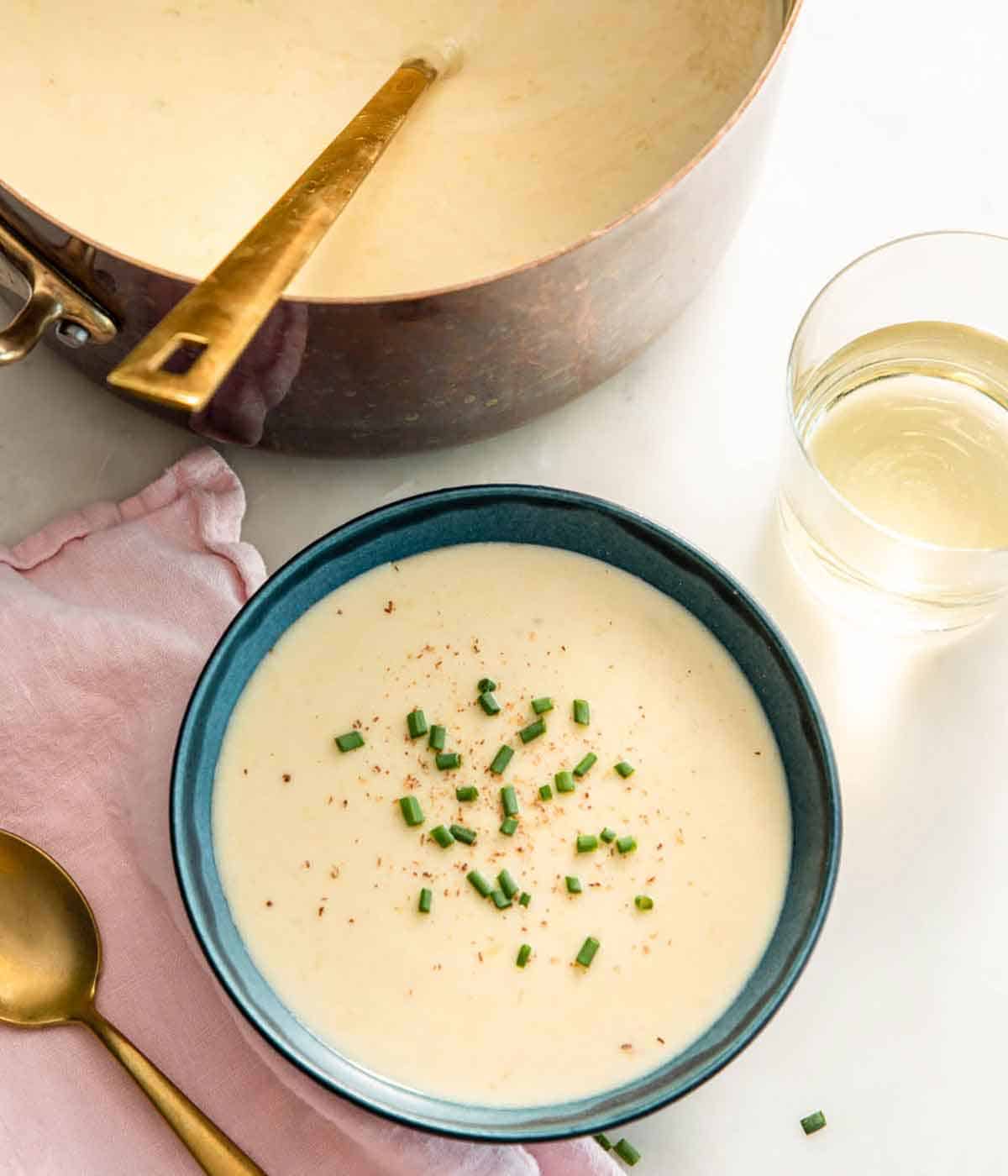 Overhead view of a bowl of vichyssoise with chives on top by a pot of more soup with a ladle.