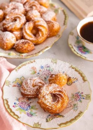 A plate of zeppole with more in the back with powdered sugar dusted on top.