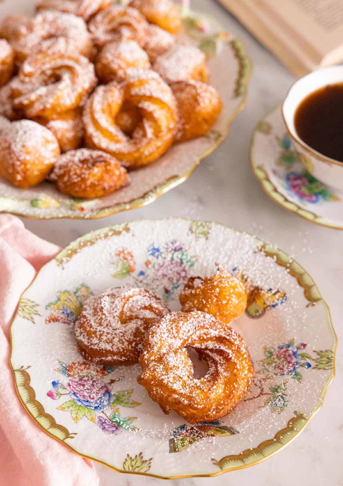 A plate of zeppole with more in the back with powdered sugar dusted on top.