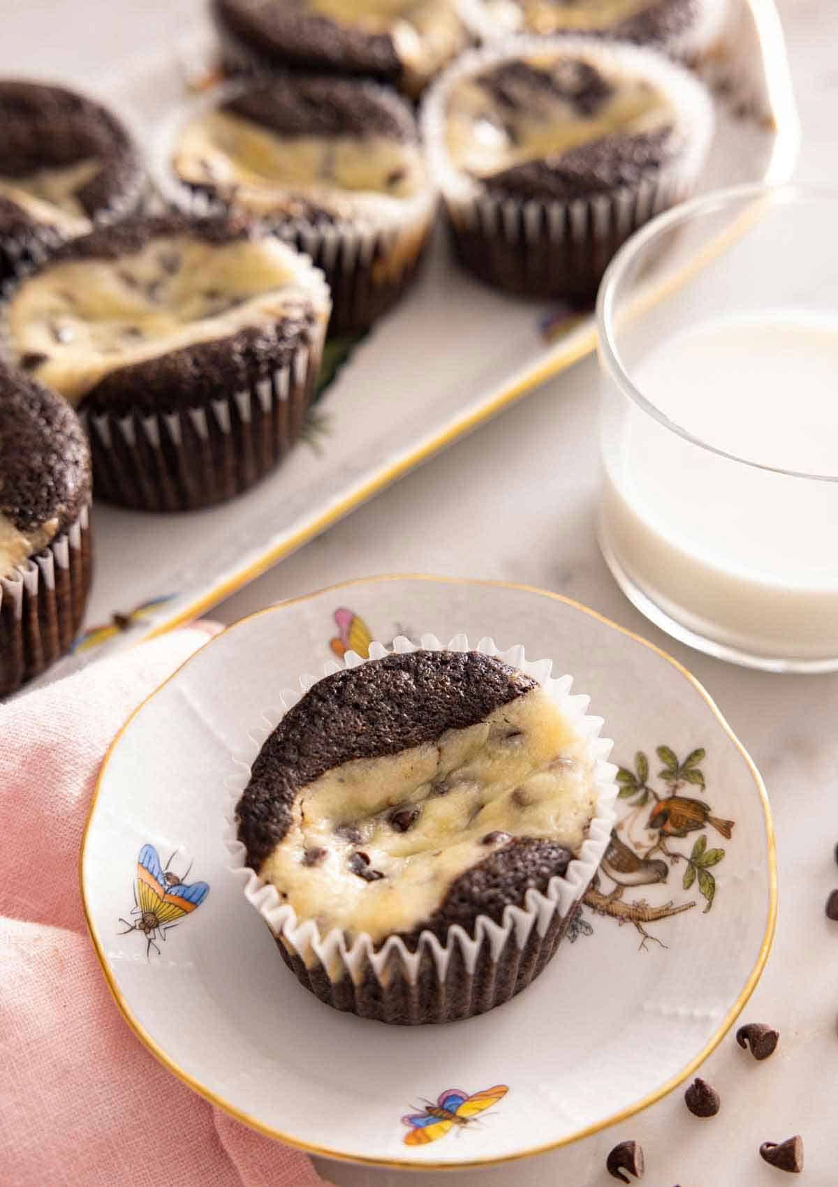 A plate with a black bottom cupcakes by a cup of milk and a platter of more cupcakes.