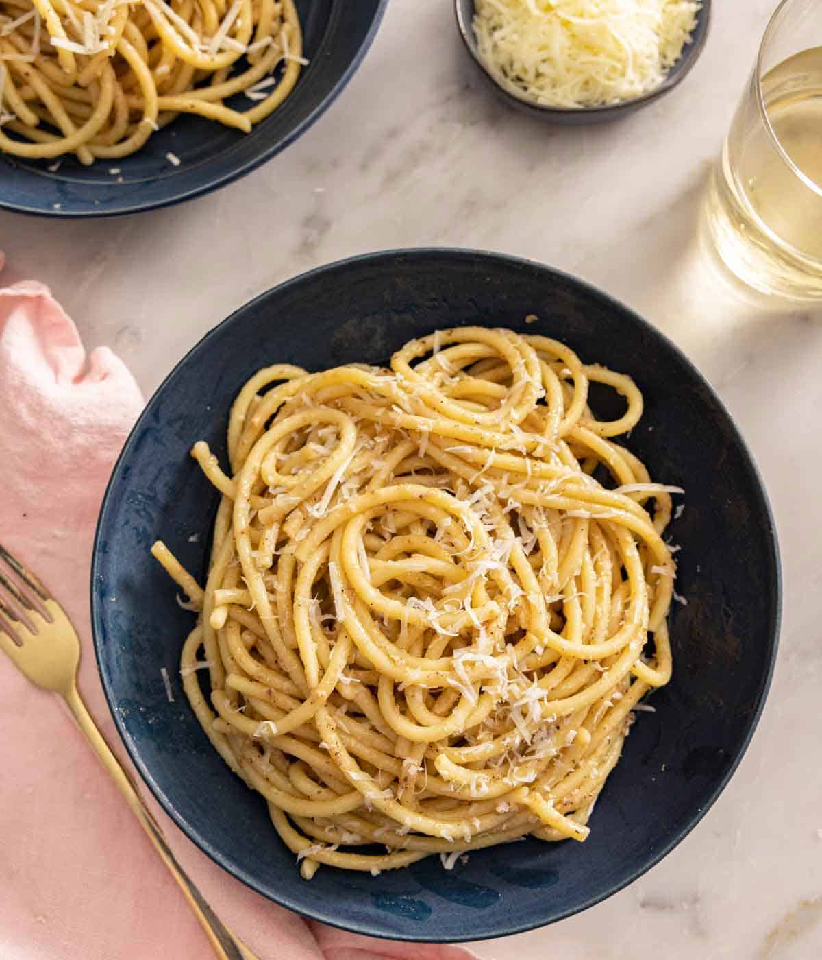 Overhead view of a plate of cacio e pepe by a fork, linen, glass of wine, and grated cheese.