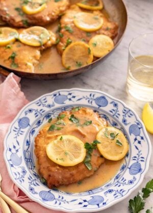 A plate of chicken Francese with a glass of wine in the background alongside a skillet.