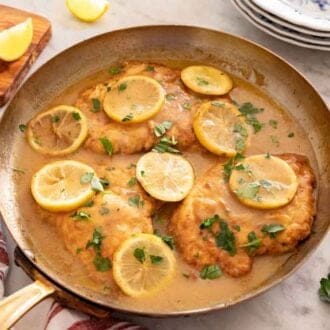 A skillet with chicken Francese with parsley and lemon on top by some plates, linen, and lemon wedges.
