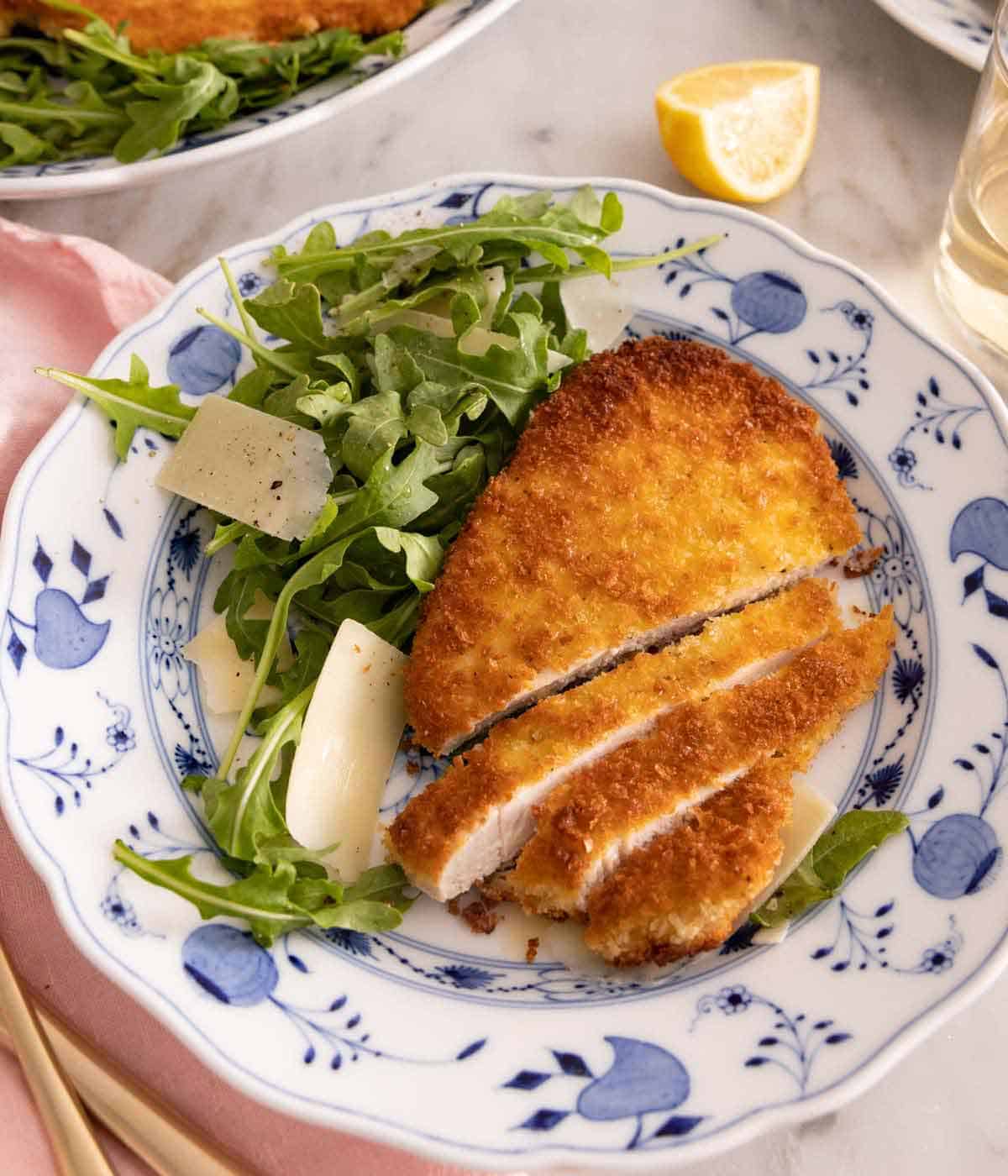 A plate with chicken Milanese with pieces sliced beside a handful of arugula with parmesan.