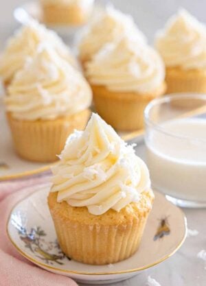 A plate with a coconut cupcake with more in the background along with milk.