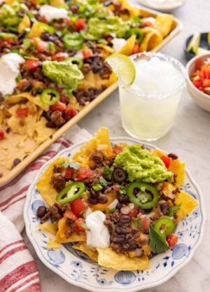 A plate with a serving of nachos by a drink and sheet pan of more nachos.