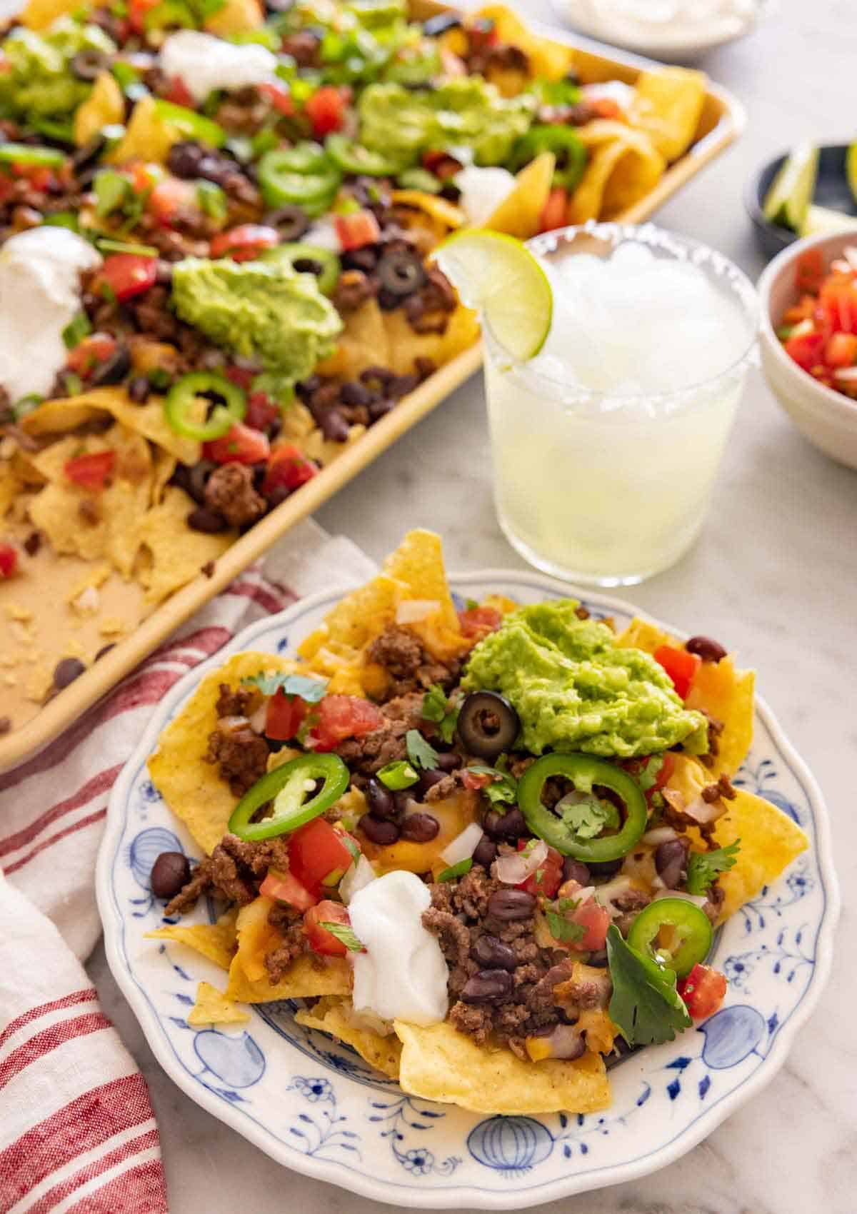 A plate with a serving of nachos by a drink and sheet pan of more nachos.
