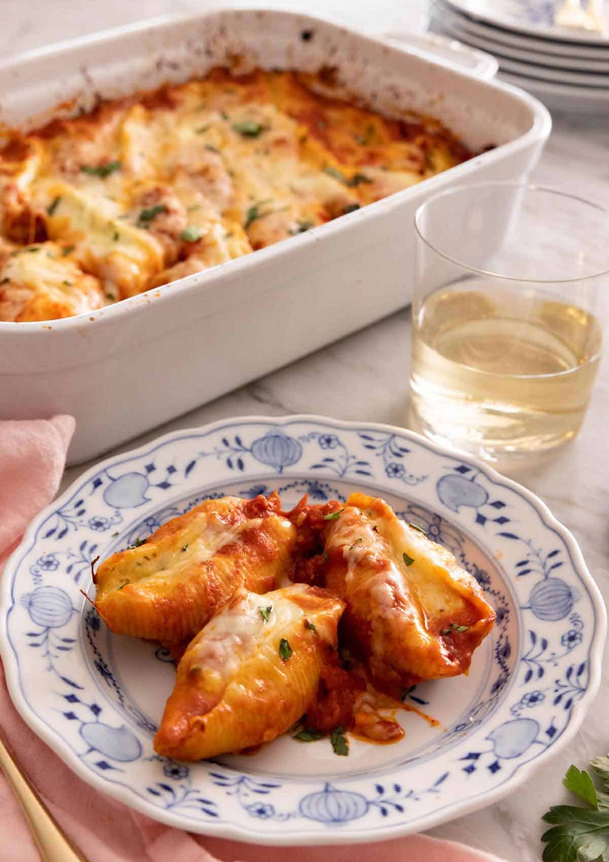 A plate with three stuffed shells by a glass of wine by a baking dish.