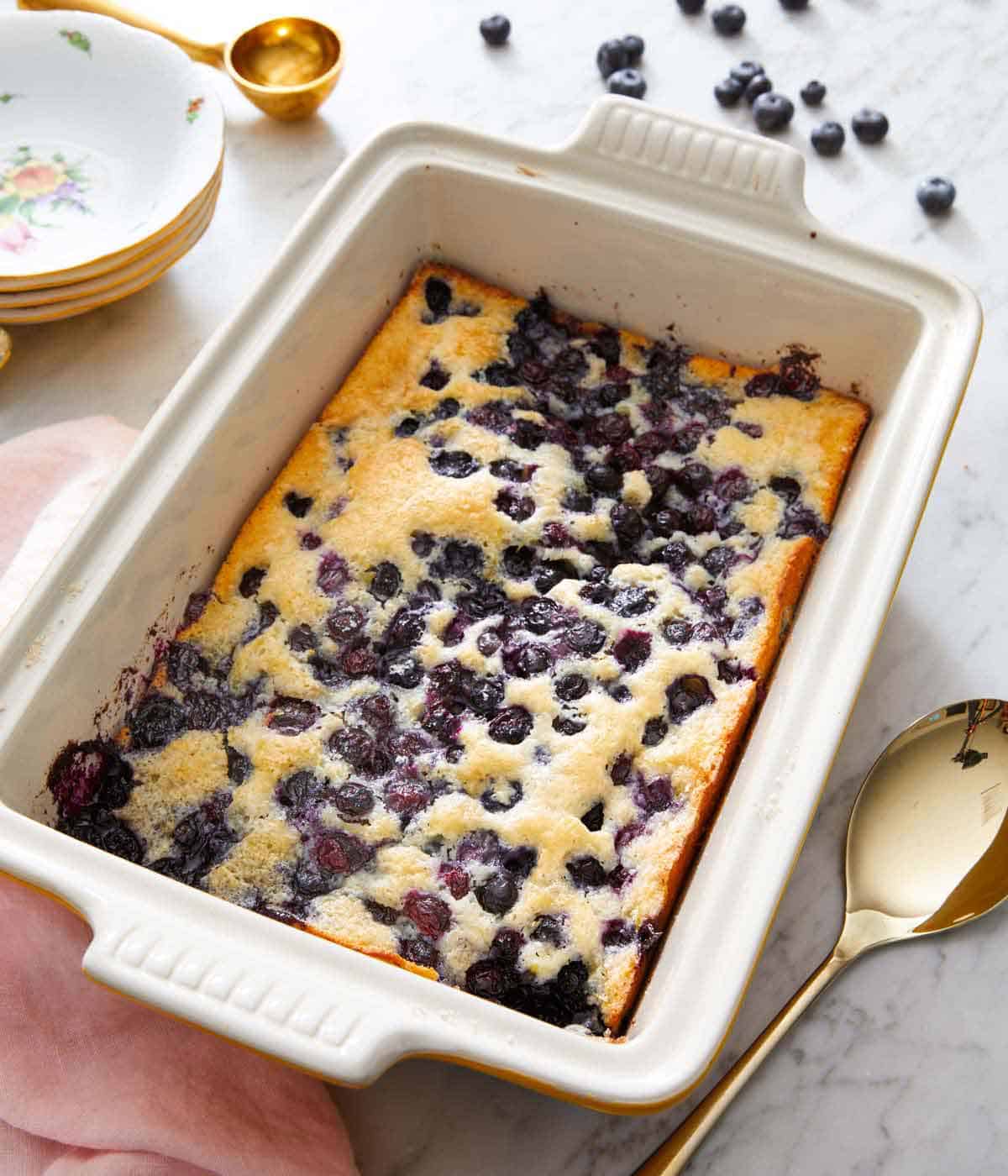 A white baking dish of blueberry cobbler with a serving spoon beside it and fresh blueberries scattered around.