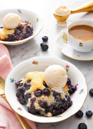 A bowl if a serving of blueberry cobbler with a scoop of vanilla ice cream on top. Mug of coffee, cobbler, and more ice cream in the background.