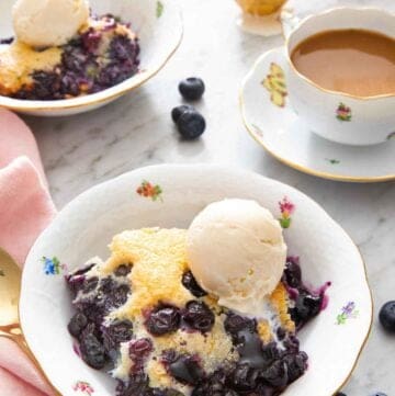 A bowl if a serving of blueberry cobbler with a scoop of vanilla ice cream on top. Mug of coffee, cobbler, and more ice cream in the background.