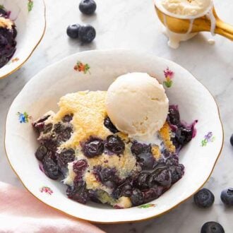 A bowl of blueberry cobbler with a scoop of ice cream on top with fresh blueberries scattered around.