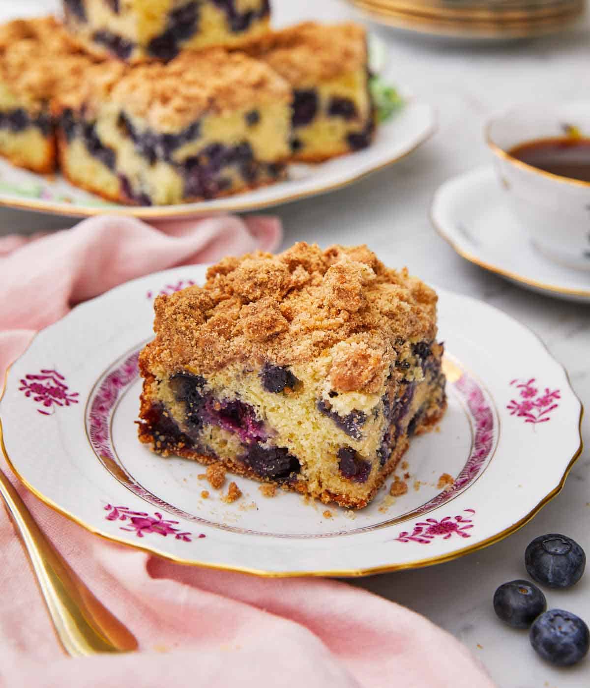 A plate with a fresh slice of blueberry coffee cake with more on a platter in the background and fresh blueberries scattered in front.