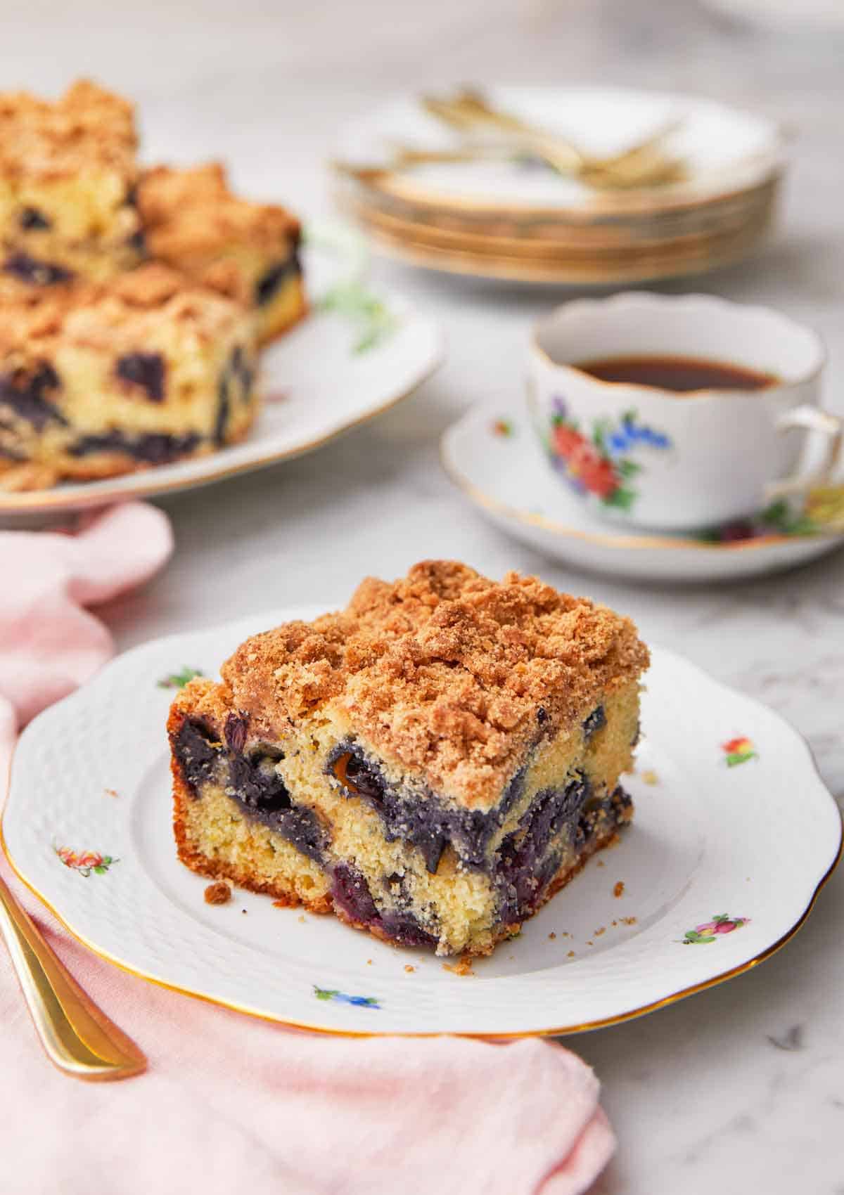 A square piece blueberry coffee cake on a plate in front of a cup of coffee.