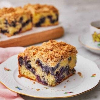 A plate with a piece of blueberry coffee cake by a cutting board with more servings and a mug of coffee.